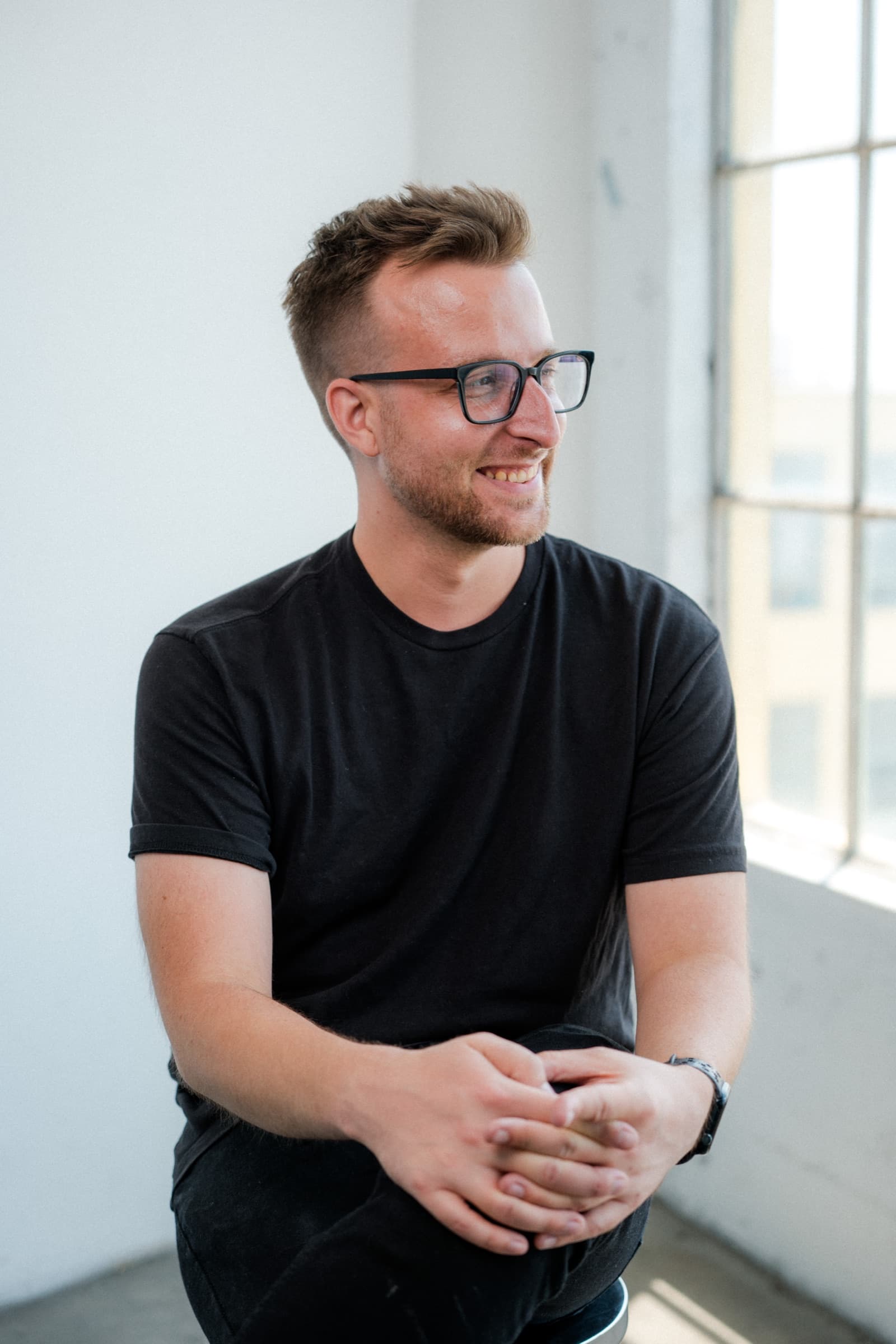 David Ryan Olson smiling in a sunlit studio, wearing glasses and a black t-shirt